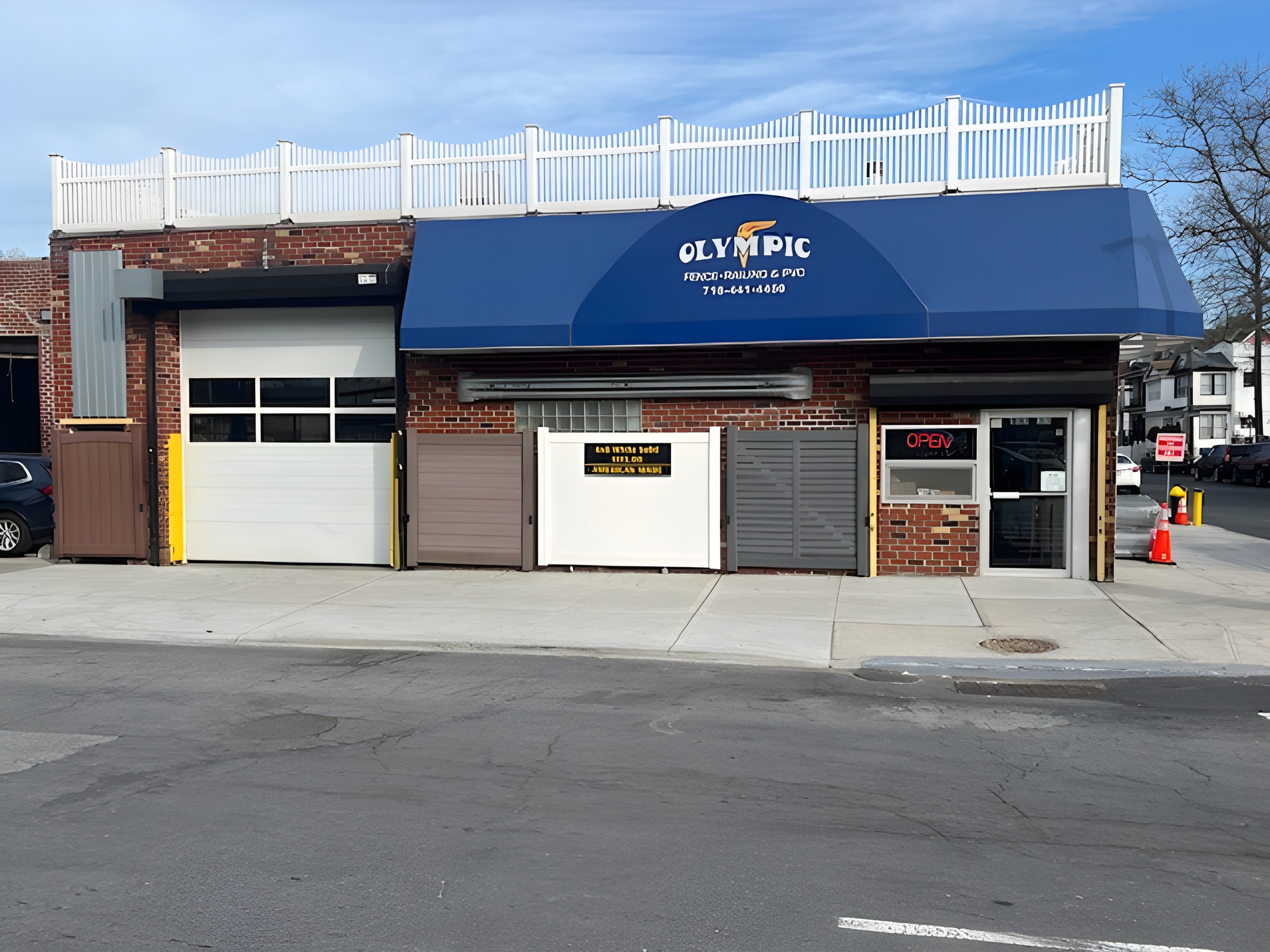 Exterior of the Olympic Fence NYC showroom and supply center in Richmond Hill, Queens, featuring fencing displays, roll-up bays, and a blue Olympic Fence awning.